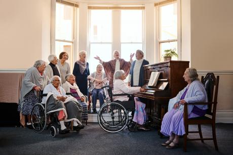 A choir of older women in front of windows, singing. Some are standing, some are seated in chairs or wheelchairs