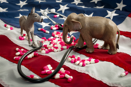 Donkey and elephant figurines face each other on a U.S. flag. A stethoscope and pills are scattered around them,