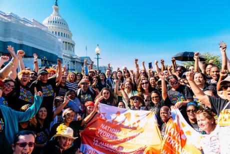 A large group of diverse people wearing National Domestic Workers Alliance shirts wave, hold banners, in front of the U.S. Capitol. Ai-jen Poo stands in the middle.