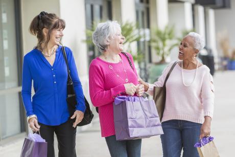 A group of three women in their 60s and 70s shopping together, carrying shopping bags, laughing and talking. They are walking on a sidewalk wearing sweaters and jeans.