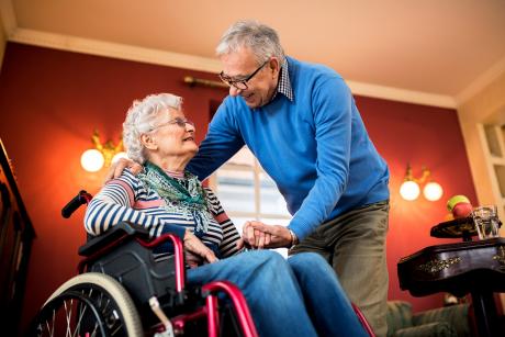 An older couple at home, the woman is using a wheelchair and the man is holding her hand and leaning down to embrace her