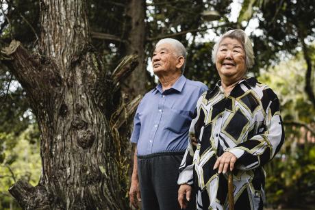 An older Japanese couple standing in front of a large tree. The man looks into the distance, the woman is smiling at the camera.