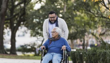 A Hispanic man with his father who is using a wheelchair in a park