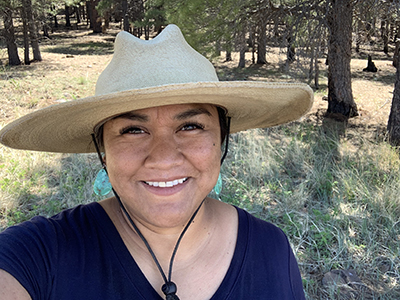 Selfie taken by Nikki Cooley. Nikki, an indigenous woman, is standing in a grove of trees, wearing a large brimmed hat and smiling at the camera.