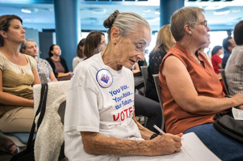 Rivko Knox sitting in a group taking notes. Her white shirt reads "Your Voice, Your Choice, Your Future" in blue and VOTE in red beneath