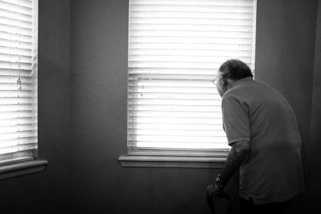 Black and white image of an older adult alone in a room, looking out the window