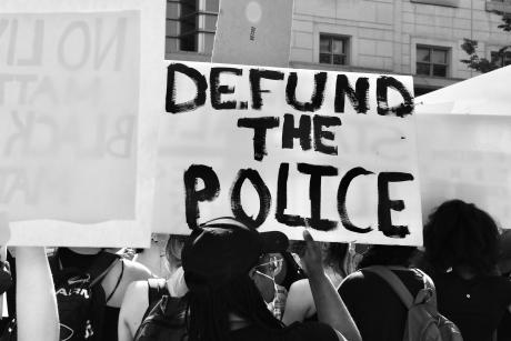 Black and white photo of a person holding sign that reads "defund the police" in a protest