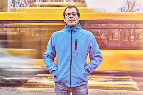 An older man in a blue jacket looking sad, a bus whizzing by behind him