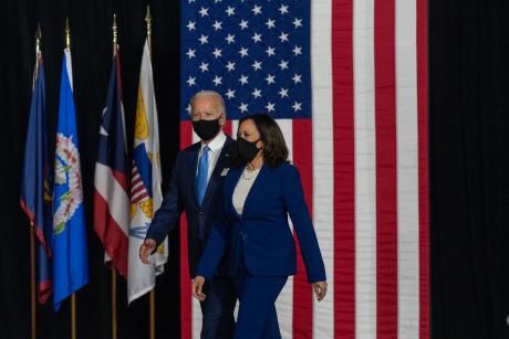 Joe Biden and Kamala Harris walk across a stage in front of US flag. Both wear masks.