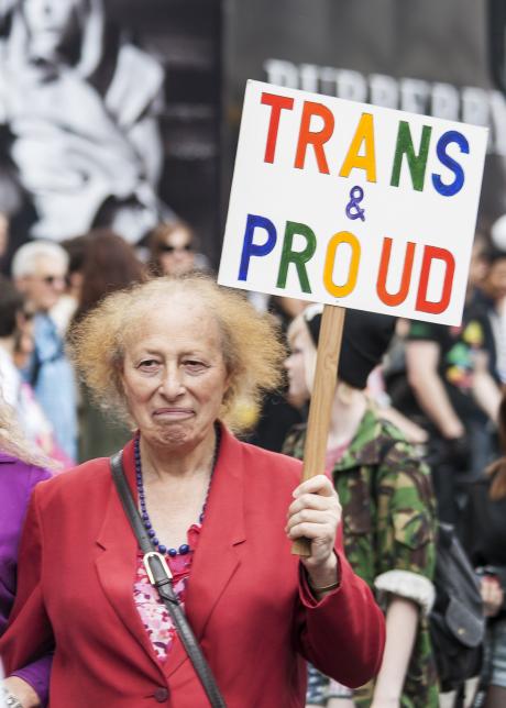 An older transgender woman holding a sign that reads "Trans & Proud"