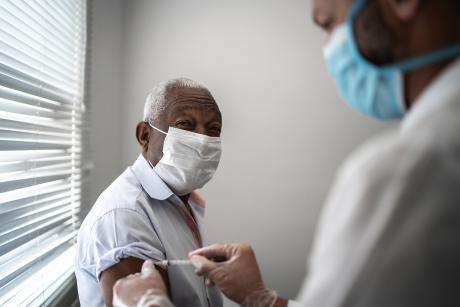An older black man wearing a mask looks at the camera as a nurse gives him a shot. His eyes are smiling behind the mask.