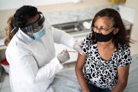 An older woman receiving a vaccine. She is wearing a black mask, the healthcare provider is wearing a mask and face shield.