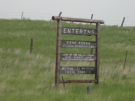 Dilapidated wooden sign the reads "Entering Pine Ridge Reservation. Land of the Ogala Siouz. Red Cloud- Black Elk - Crazy Horse. No hunting without tribal permits."
