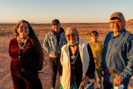 An Indigenous family outside, smiling at the camera. Family includes two middle-aged people, one older woman, a teenager and a youth.