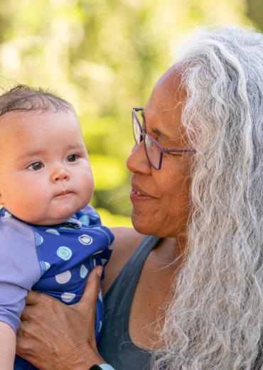 Older Indigenous woman with long gray hair holds baby
